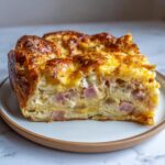 A close-up slice of rich Croissant Breakfast Casserole showing layers of flaky pastry, ham, and cheesy egg filling.