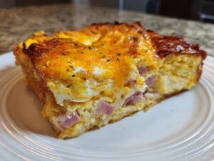 A close-up of a rich slice of Croissant Breakfast Casserole featuring baked egg, ham cubes, and melted cheddar cheese.