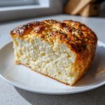 A thick slice of fluffy cottage cheese bread, golden brown on top and sprinkled with black seeds, resting on a white plate.