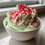 Close-up of fluffy, light green grinch dip topped generously with bright red sprinkles in a white bowl.