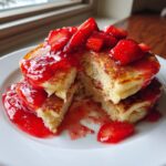 A stack of fluffy strawberry pancakes cut open, generously topped with bright red strawberry sauce and fresh sliced strawberries.