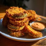 A stack of perfectly golden, crispy fried green tomatoes served on a plate next to a small bowl of creamy dipping sauce.