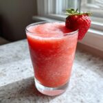 A vibrant, frozen strawberry daiquiri garnished with a fresh strawberry, sitting on a speckled countertop near a window.
