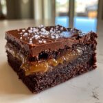 Close-up of a fudgy caramel brownie slice with a layer of gooey caramel and topped with chocolate ganache and flaky sea salt.