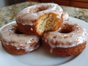 Close-up of three glazed cake donuts stacked on a white plate, one having a bite taken out showing the crumb.