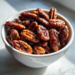 A white bowl filled with shiny, dark brown candied pecans, glistening under bright light.