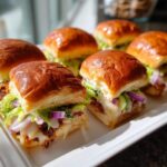Close-up of several freshly baked grinder sliders with melted cheese, lettuce, and red onion on a white serving tray.