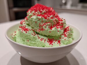 Close-up of bright green Grinch dip mounded high in a white bowl and covered in red nonpareil sprinkles.
