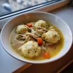 A close-up of a white bowl filled with rich chicken and dumplings soup, featuring shredded chicken, carrots, and herbs.