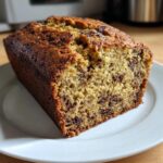 A close-up of a freshly baked, moist banana bread recipe loaf with visible chocolate chips on a white plate.