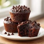 Three moist chocolate muffins stacked on a white plate, one with a bite taken out, topped with chocolate chips.