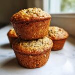 A stack of two golden brown, moist oatmeal muffins with visible oats on top, with more muffins blurred in the background.