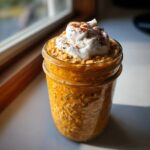 Close-up of bright orange pumpkin overnight oats in a mason jar, topped with whipped cream and cinnamon.