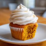 A single pumpkin spice cupcake with a bite taken out, topped with thick swirls of white cream cheese frosting.