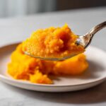 A spoonful of bright orange, thick homemade pumpkin puree being lifted over a white plate.