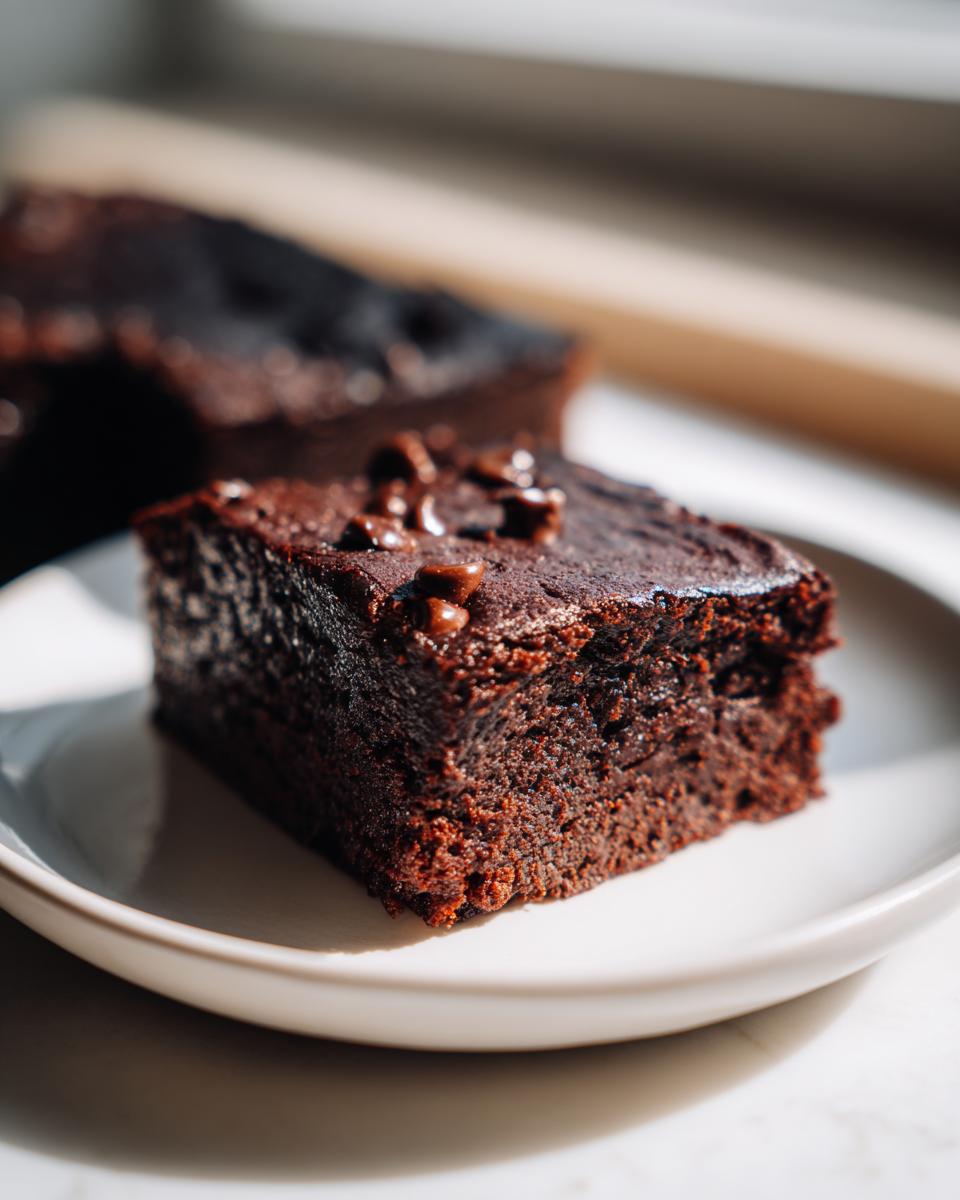 Close-up of a rich, dark vegan brownies square with a shiny top and chocolate chips, served on a white plate.