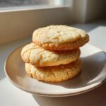 Three soft sugar cookies stacked on a small speckled plate, highlighted by bright sunlight.