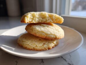 Three soft sugar cookies stacked on a white plate, with the top one broken to show the fluffy interior.