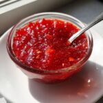 A close-up of bright red, chunky strawberry freezer jam served in a small clear glass bowl with a spoon resting inside.
