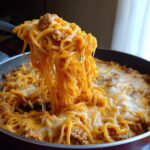 A serving spoon lifts a portion of cheesy taco pasta, showing long, melted cheese strings stretching from the pan.