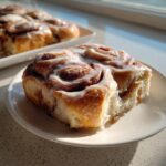 A single, gooey tiktok cinnamon roll covered in white icing sits on a small white plate, with more rolls blurred in the background.
