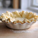Close-up of an unbaked, flaky pie crust dough perfectly crimped in a white ceramic pie dish.