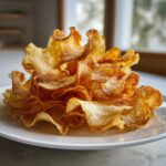 A mound of golden, wavy homemade potato chips seasoned with salt piled high on a white plate.