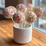 A bouquet of white chocolate cake pops covered in colorful sprinkles displayed in a white ceramic holder.