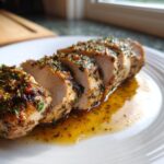 Close-up of a perfectly cooked and sliced pork tenderloin recipe resting in its own juices on a white plate.