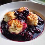 Three golden-topped blackberry dumplings swimming in a rich, dark purple blackberry sauce.