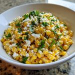 A close-up of a white bowl filled with creamy, bright yellow street corn salad topped with crumbled white cheese and fresh cilantro.