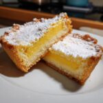 Two halves of a baby lemon impossible pie showing the bright yellow filling and shortbread crust, dusted with powdered sugar.