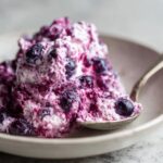A close-up of creamy, purple-swirled blueberry fluff salad in a light bowl with a silver spoon scooping some out.