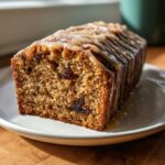 A slice of moist cardamom date cake topped with a thick caramel glaze, sitting on a white plate.
