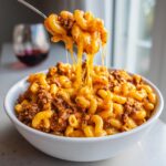 A fork lifting a bite of cheesy cheeseburger pasta, showing long, stretchy cheese pulls over a bowl of macaroni and ground beef.