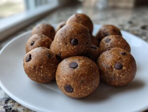 A pile of homemade, round chocolate chip protein balls resting on a white plate near a window.