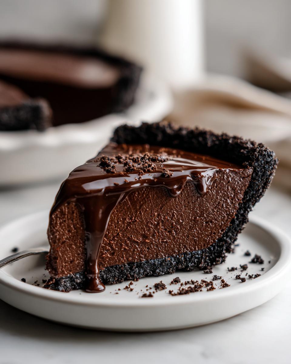 A decadent slice of chocolate fudge pie with a dark cookie crust, topped with glossy chocolate ganache.