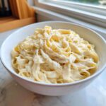 A close-up of fettuccine pasta coated in rich, creamy Olive Garden Alfredo Sauce, topped with grated cheese and pepper.