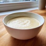 A close-up of a creamy, pale yellow bowl of amazing garlic soup, lightly seasoned with pepper.