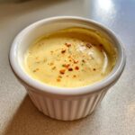 Close-up of creamy, yellow spicy caesar dressing topped with red pepper flakes in a small white ramekin.