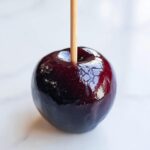 Close-up of a single, perfectly dipped dark jolly rancher candy apple with a wooden stick inserted, resting on a white surface.