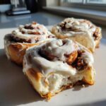 Three gooey easy homemade cinnamon rolls topped with thick white frosting, one has a bite taken out.