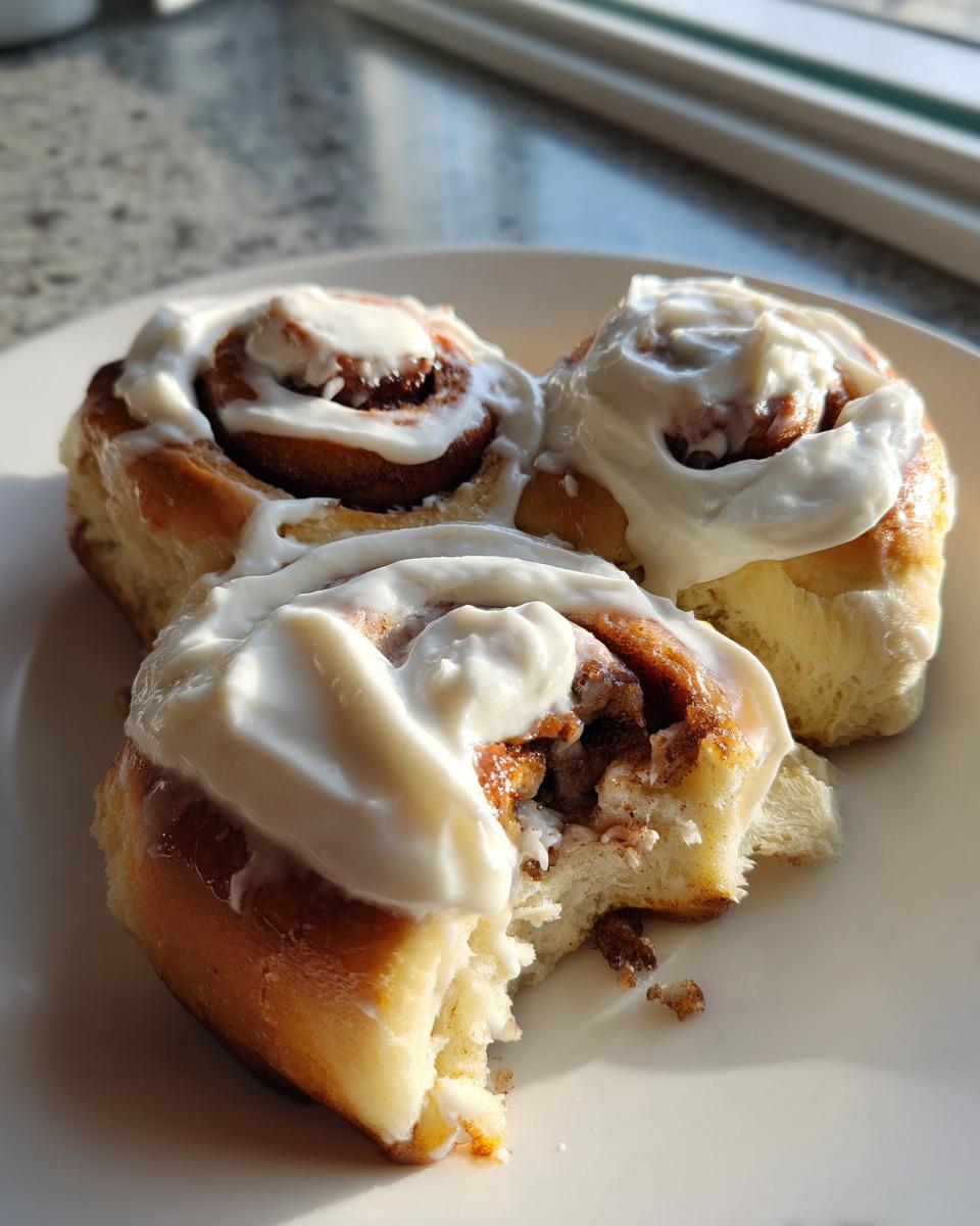 Three soft easy homemade cinnamon rolls generously topped with white cream cheese frosting on a white plate.