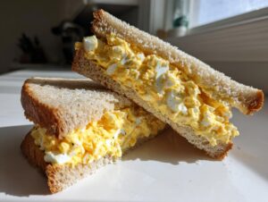 A close-up of a delicious egg salad sandwich, cut in half diagonally, showing creamy yellow filling on whole wheat bread.