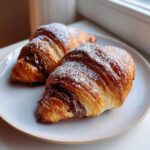 Two golden brown chocolate croissant pastries, oozing melted chocolate and dusted with powdered sugar, served on a light plate.