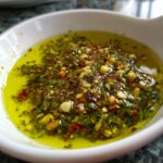 Close-up of a vibrant olive oil bread dip mixture with green herbs, chopped garlic, and red pepper flakes.