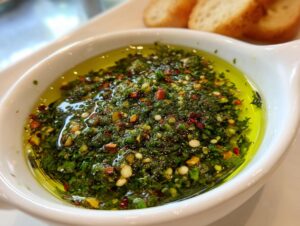Close-up of a white bowl filled with vibrant green olive oil bread dip, featuring herbs, spices, and chili flakes.