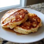 Two golden-brown, fluffy muffin mix pancakes stacked slightly on a white plate, drizzled with syrup.