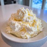 A large, fluffy mound of pale yellow vanilla buttercream frosting piled high on a white plate.