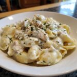 A close-up of a white bowl filled with creamy garlic butter chicken alfredo shells pasta, topped with fresh parsley.